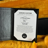 A Gibson Custom Certificate of Authenticity card displayed inside an open black folder on a bright yellow-orange furry surface. The certificate includes the maker name Gibson and dealer details.