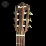 Close-up of the headstock of an Ibanez classical guitar against a solid black background, showing the brand name 'Ibanez' in white on the dark wood headstock, six tuning pegs, and tuning machines.
