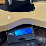 Close-up of a cream-colored electric guitar body resting on a digital smart weigh scale displaying 7.6 kg, set on a dark wooden surface.
