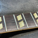 Close-up of a guitar fretboard with dark wood blocks, silver metal fret edges, and pale cream inlays on a dark textured surface.