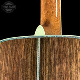 Close-up detail of an acoustic guitar body showing the rich wood grain, a decorative inlay line, and part of the neck against a black background. A Tone Shop watermark is visible in the top left corner.