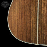 Close-up detail of an acoustic guitar body showing a rich brown wood grain finish with a silver-toned binding edge against a black background. A Tone Shop watermark is visible in the top-left corner.