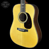 A close-up studio shot of a wooden acoustic guitar against a solid black background, featuring a natural wood top, dark brown sides, and a black pickguard.