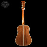 The back of an acoustic guitar with a rich reddish-brown wood finish and gold tuning keys, shown against a solid black background with a Tone Shop watermark in the top left corner.