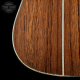 Close-up of an acoustic guitar body showing a rich brown wood grain finish with a decorative inlay running vertically and along the lower edge, set against a black background with a Tone Shop watermark in the top left.