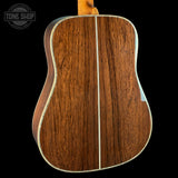 Close-up of the back of an acoustic guitar showing a rich brown wood grain finish with a light cream binding, set against a solid black background.