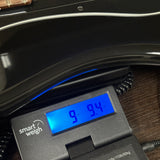 A close-up of a black digital smart weigh scale displaying 9.94 kg under a glossy black guitar body on a dark wooden surface.