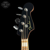 Close-up of a Fender bass guitar headstock in black with gold script branding, four silver tuning pegs, and part of the light wood fretboard visible against a black background.