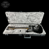 A black electric guitar with a gold fretboard and silver hardware, displayed inside an open gray hard case with a white textured interior. The Tone Shop logo is visible in the top left corner.