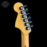Close-up of the headstock of a Fender Stratocaster electric guitar, showing the natural maple wood finish and black tuning pegs with blue-white labels, set against a solid black background.
