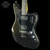 A black electric guitar with a glossy finish, featuring a black pickguard, chrome hardware, and a yellow headstock visible at the top, set against a solid black background with a Tone Shop watermark in the upper left corner.