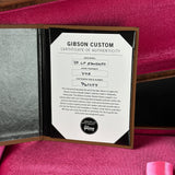 An open Gibson Custom Certificate of Authenticity booklet displayed on a bright pink velvet surface. The white certificate is centered inside a black-lined folder with a brown border, showing model and serial details for a '57 LP Standard.
