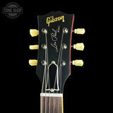 Close-up of a Gibson Les Paul guitar headstock, featuring a black finish, gold Gibson logo, silver script signature, and cream-colored tuning pegs against a solid black background.