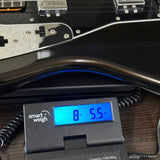 A close-up of a black electric guitar body resting on a digital smart weigh scale displaying 8.55 kg, set on a dark wooden surface.