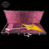 A yellow electric guitar resting inside an open black hard case with a textured magenta-pink interior, shown against a black background. A Tone Shop logo watermark is visible in the top-left corner.
