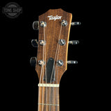 Close-up of a Taylor acoustic guitar headstock in dark brown wood with six silver tuning pegs and six strings, shown against a solid black background with a Tone Shop watermark in the top left corner.