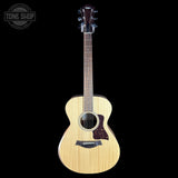 A Taylor acoustic guitar with a natural wood finish and dark pickguard, shown against a solid black background with a Tone Shop watermark in the top left corner.