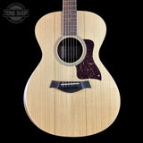 A Taylor acoustic guitar with a natural flamed maple top, dark brown pickguard, and black bridge, shown against a solid black background with a Tone Shop watermark in the top left corner.