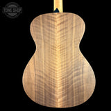 Close-up of the back of a guitar body made from figured wood, showing a prominent natural grain pattern in warm tan and brown tones against a solid black background. A Tone Shop watermark is visible in the top-left corner.