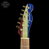 Close-up of a Fender electric guitar headstock featuring a vertical glitter stripe design in red, gold, and blue, with gold tuning pegs and a dark background. A Tone Shop watermark is visible in the top-left corner.