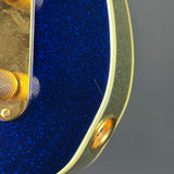Close-up of an electric guitar body featuring a deep blue glitter finish, gold hardware, and a cream-colored binding on a gray background.