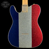 A close-up of the back of a single-cutaway electric guitar with a glittery French flag design: vertical stripes in blue, silver, and red, with a light wood neck visible at the top against a black background.