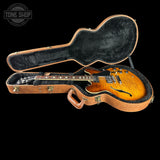 A sunburst electric guitar resting inside an open brown hard case with black plush lining, shown against a solid black background. A Tone Shop watermark is visible in the top-left corner.