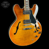 A double-cutaway electric guitar with a natural flame maple top, featuring a warm orange-brown sunburst finish, black pickguard, chrome hardware, and a dark fretboard. The guitar is photographed against a solid black background with a Tone Shop logo watermark in the top-left corner.