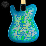 A blue electric guitar body with a vibrant, all-over retro floral pattern in pink, green, and light blue, shown against a solid black background. A Tone Shop logo is visible in the top-left corner.