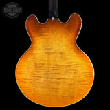 The back of an electric guitar with a natural flame maple wood finish, showing a warm honey-orange color and prominent wood grain, set against a solid black background.