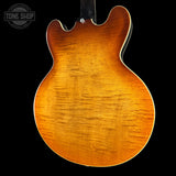 Close-up of the back of an electric guitar body showing a flamed maple wood finish in a warm orange-brown sunburst, photographed against a black background with a Tone Shop watermark in the upper left.