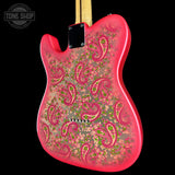 A close-up of a guitar body back showing a vibrant pink outer edge and a detailed paisley floral pattern in pink, green, and gold over a dark base. A black plate and a light wood neck are visible at the top against a solid black background.