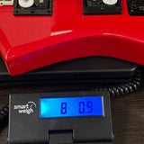 A close-up of a red electric bass guitar body with a black coiled cable connected to a black Smart Weigh electronic scale displaying weight on its blue backlit screen.