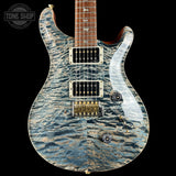 A close-up of a PRS electric guitar body featuring a distinctive blue and gray wavy quilted maple top, with a dark brown fretboard and gold hardware, set against a solid black background.