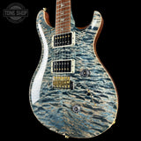 A close-up of an electric guitar body with a blue and gray flamed maple wood finish, featuring gold hardware and a dark brown fretboard, shown against a black background with a Tone Shop watermark in the top-left corner.
