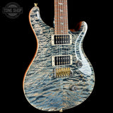 A close-up of a double-cutaway electric guitar featuring a blue and gray flamed maple top with a wavy, textured finish. The guitar has a dark brown fretboard, gold hardware, and is photographed against a solid black background.