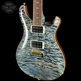 A close-up of an electric guitar featuring a blue and gray flamed maple top with a wavy, textured finish. The guitar has a dark brown fingerboard, gold hardware, and white hardware accents, shown against a solid black background with a Tone Shop watermark in the top-left corner.