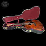 An acoustic guitar with a warm brown wooden body and dark neck, displayed inside an open black hard case with a burgundy velvet lining, set against a solid black background.