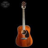 A wooden acoustic guitar with a warm reddish-brown finish and a black pickguard, shown vertically against a solid black background. A circular 'TONE SHOP' watermark is visible in the top-left corner.