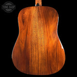 The back of an acoustic guitar made of rich, warm-toned wood, showing detailed grain patterns, set against a solid black background with a Tone Shop watermark in the top left corner.