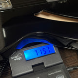A close-up of a smart weigh electronic scale displaying a weight of 715.1 kg on its blue backlit screen, positioned under a black guitar body on a wooden surface.
