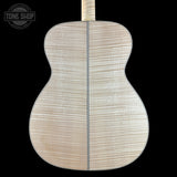 The back of an acoustic guitar made of figured maple wood, showing a light beige natural finish with prominent grain patterns and a dark center seam, set against a solid black background.