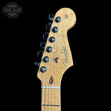 Close-up of a Fender Stratocaster electric guitar headstock in a natural maple finish, showing six black tuning pegs and the Fender logo, set against a solid black background.