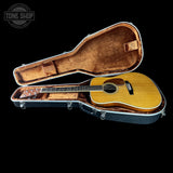 An acoustic guitar with a natural wood finish displayed inside an open black hard case with brown velvet lining, set against a solid black background. A Tone Shop watermark is visible in the top-left corner.