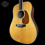 A close-up studio shot of an acoustic guitar body against a black background, showing a natural wood finish, sound hole, pickguard, and part of the neck. A Tone Shop watermark is visible in the upper left corner.