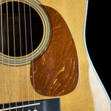 Close-up of an acoustic guitar body showing the natural wood soundboard, circular sound hole with decorative rosette, strings, pickguard, and part of the black headstock.