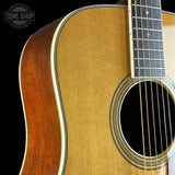 Close-up of an acoustic guitar body showing the natural wood grain top, sound hole rosette, and part of the dark fretboard, with a Tone Shop watermark in the upper left corner.
