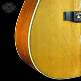 Close-up detail of an acoustic guitar body showing the natural wood side edge, the yellow-gold tonewood top, a black bridge, and a white binding line against a black background. A Tone Shop watermark is visible in the upper left.