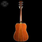 The back of a wooden acoustic guitar, showing the rich wood grain on the soundboard and neck, photographed against a solid black background with a Tone Shop watermark in the top left corner.