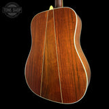 Close-up of the back of an acoustic guitar showing a rich reddish-brown wood grain finish with white binding, set against a solid black background. A Tone Shop watermark is visible in the top-left corner.